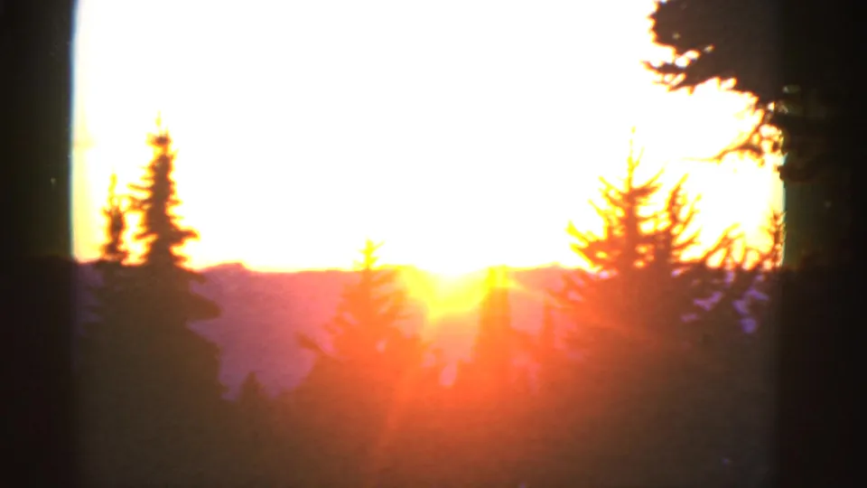 Low golden sun flaring over distant mountains, framed by evergreen silhouettes in vintage 8mm footage.