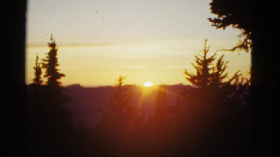 Soft 1950s 8mm view of sunlit mountain ridges and dark evergreen silhouettes, with warm exposure and film grain.