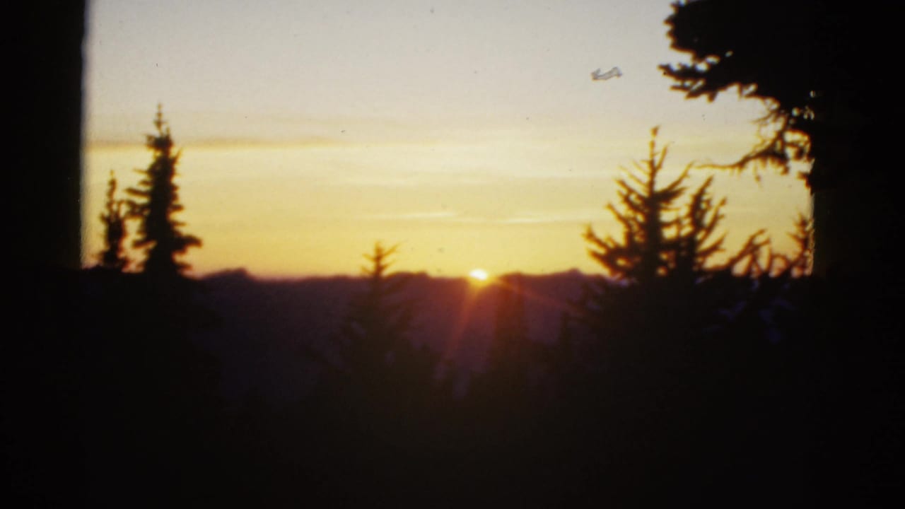 Sunrise over a distant ridge in archival 8mm footage, with evergreen silhouettes and a small aircraft shape high in the sky.