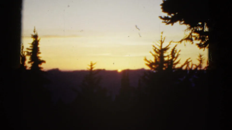 1950s 8mm home-movie frame showing a golden sunset beyond silhouetted evergreens and dark mountain ridges.