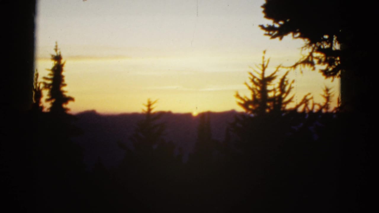 1950s 8mm frame with the sun dropping behind distant ridges, framed by dark evergreen trees.