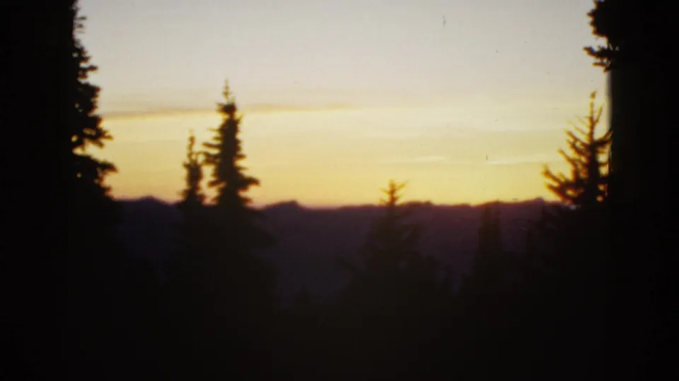 Early-1950s 8mm frame showing pine outlines, a distant ridge line, and a bright band of sunset sky.