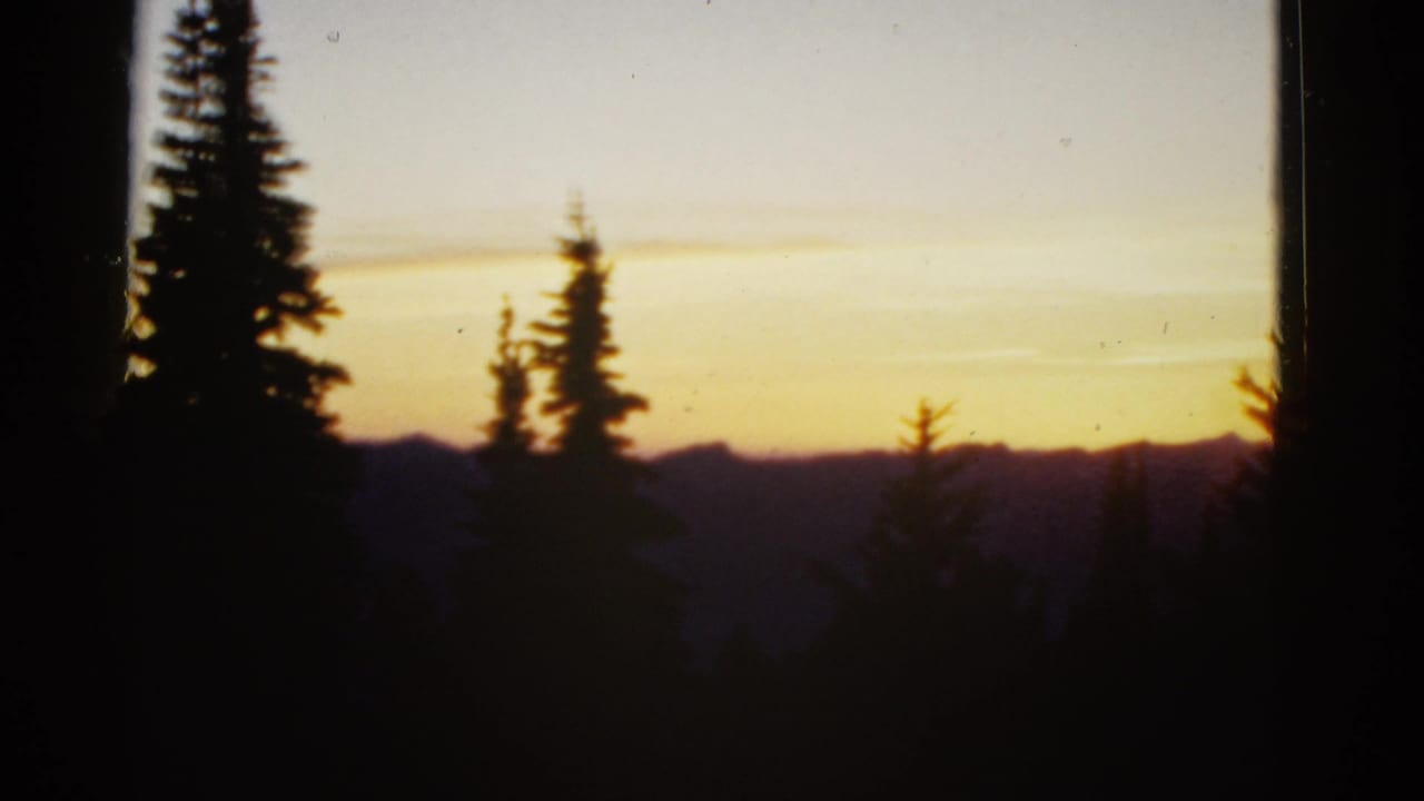 1950s 8mm home-movie frame showing dark evergreen trees before a golden sunset and shadowed mountain horizon.