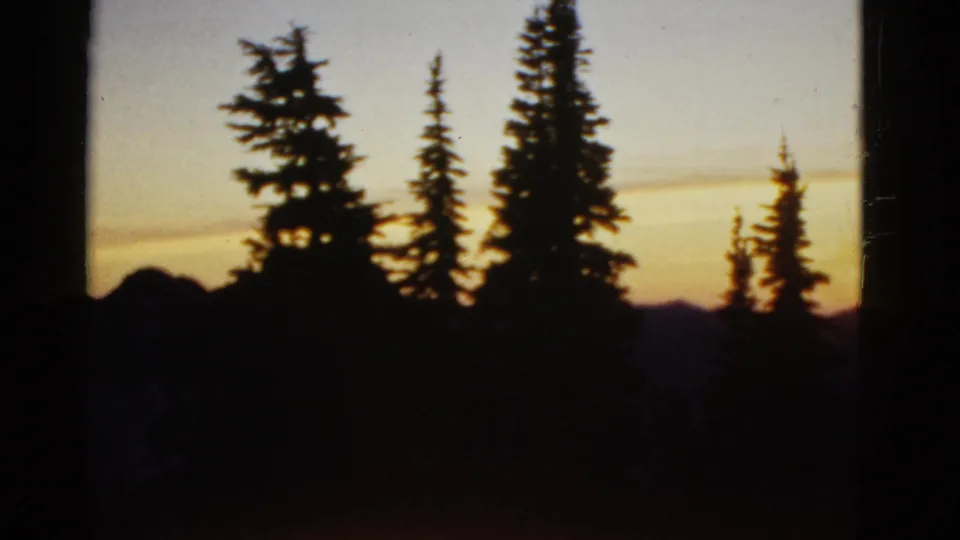 1950s 8mm frame showing evergreen silhouettes, a dark lower foreground, and warm sunset color over a mountain horizon.