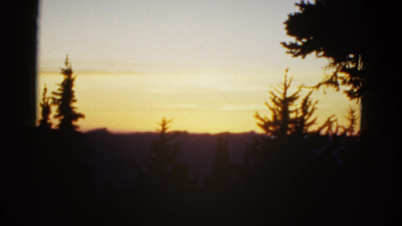 Early-1950s 8mm frame of a golden sunset over distant hills, with evergreen silhouettes in the foreground.