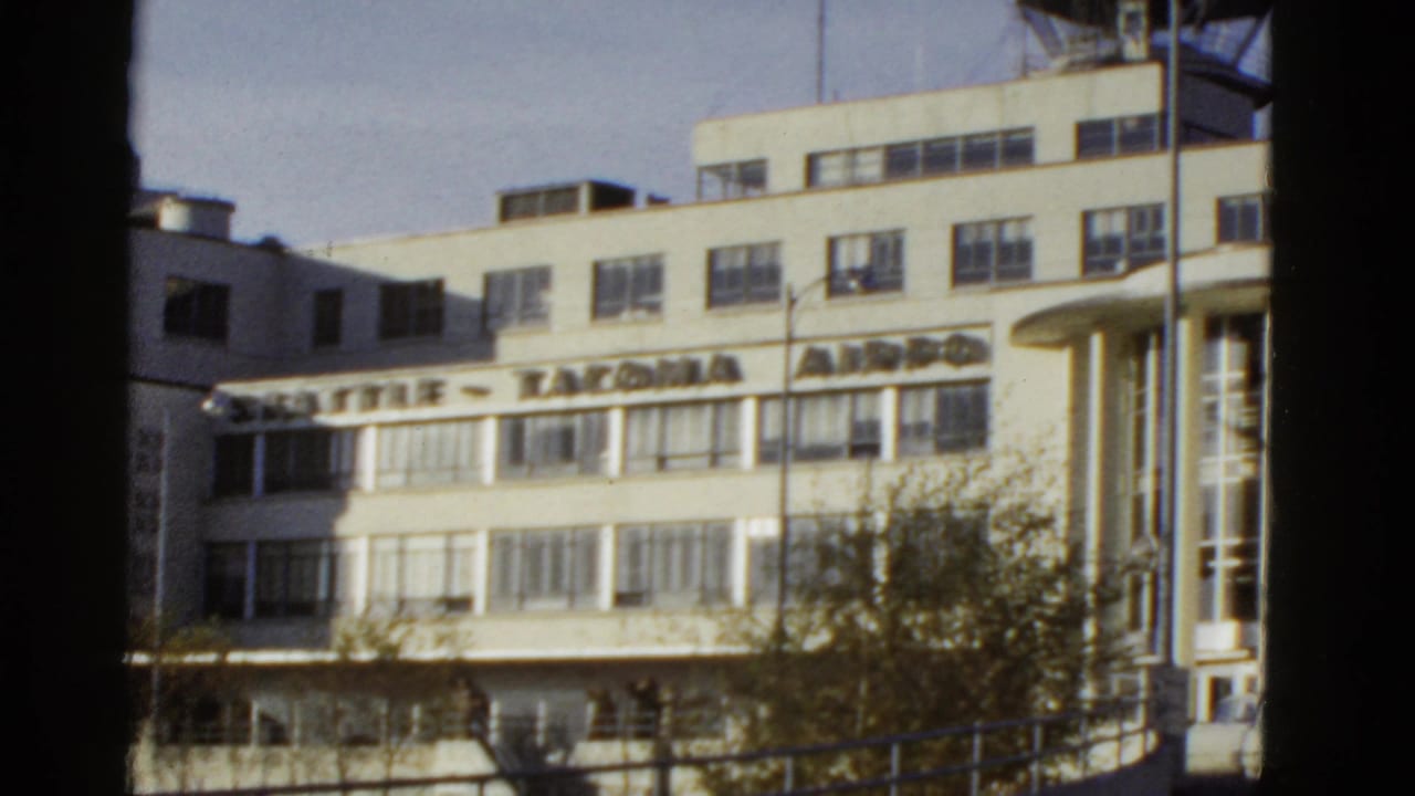 Exterior view of a mid-century airport terminal with visible Seattle-Tacoma Airport signage, pale facade, windows, and trees in front.