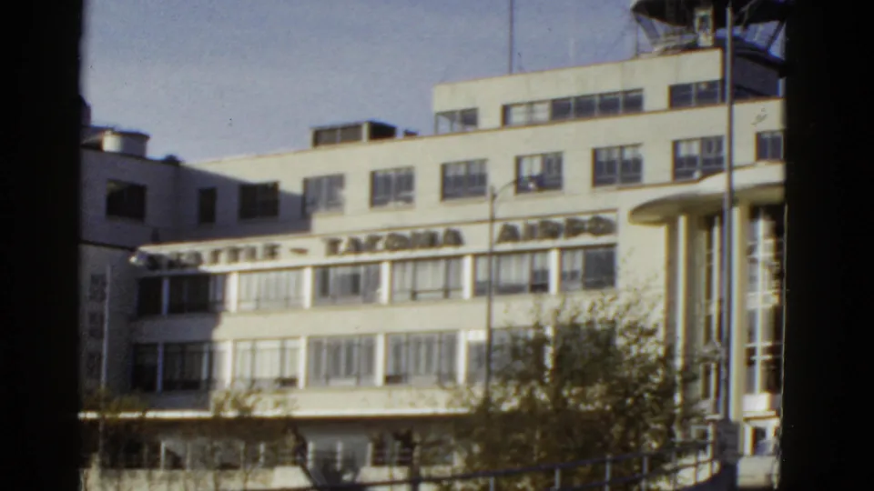 Handheld 8mm view of the Seattle-Tacoma Airport terminal facade, showing rooftop lettering, horizontal windows, and a tree-lined foreground.
