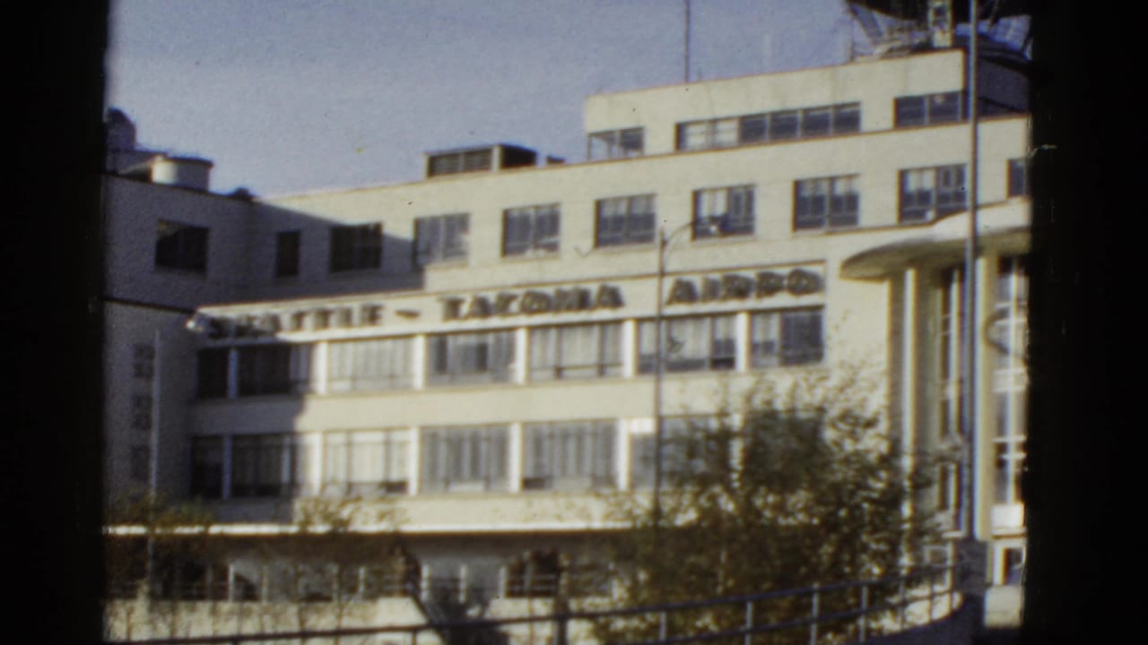 Reported 1950s 8mm view of an airport terminal exterior with white facade, rows of windows, large signage and blue sky.