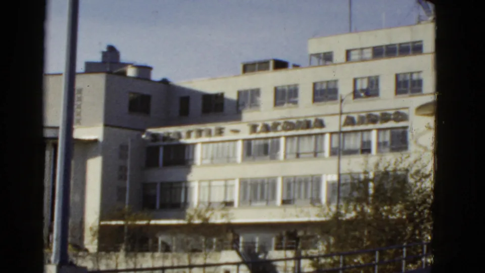 Handheld 8mm view of a white airport terminal facade, with large signage partly visible above window rows and a pole at left.