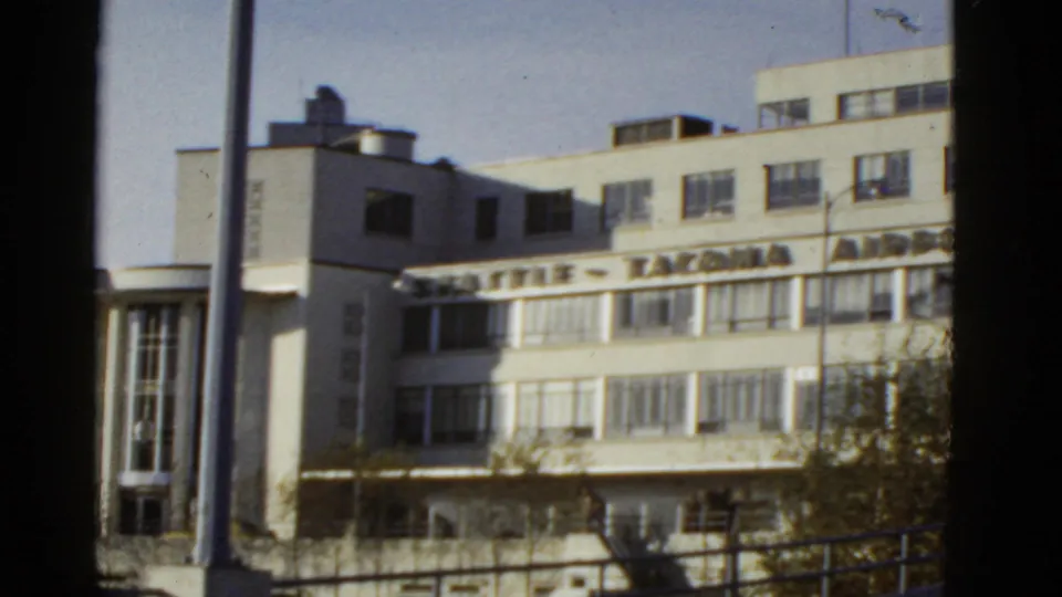 Reported 1950s 8mm frame of an airport terminal exterior, showing signage across a pale facade with trees and railings below.
