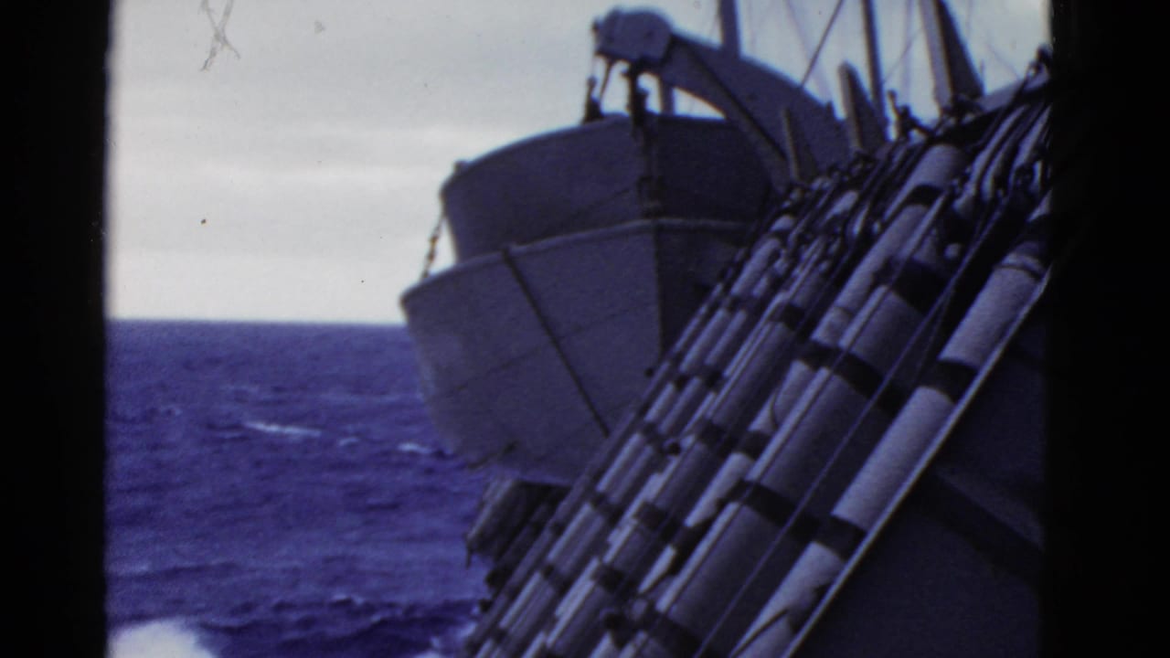 Handheld 8mm shipboard view showing the vessel side, lifeboat gear, railings, and blue ocean horizon.