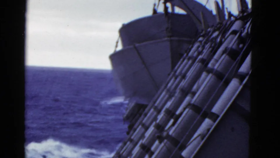 1950s 8mm shipboard frame with railings and lifeboat gear tilted over blue water and white wake.