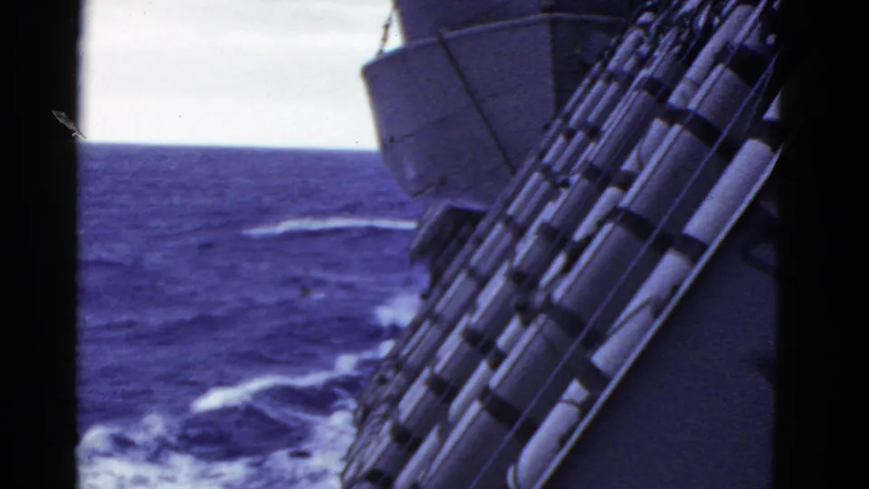 Handheld 8mm view from a ship, with deck railings and lifeboat equipment beside choppy blue ocean.