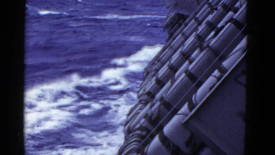Vintage 8mm view from a moving ship, with foamy water below and cylindrical fittings along the side.