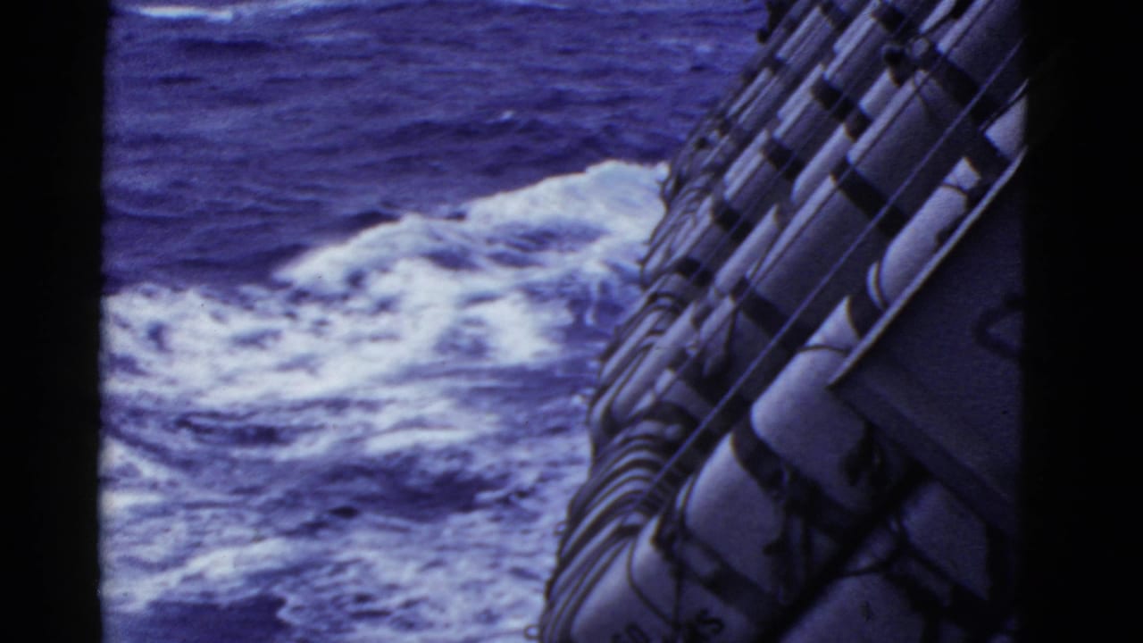 1950s 8mm home-movie frame looking past secured life rafts toward churning ocean water near the side of a moving vessel.