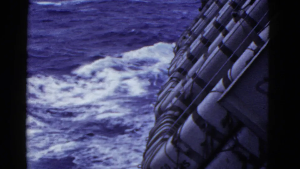 1950s 8mm home-movie frame looking past secured life rafts toward churning ocean water near the side of a moving vessel.