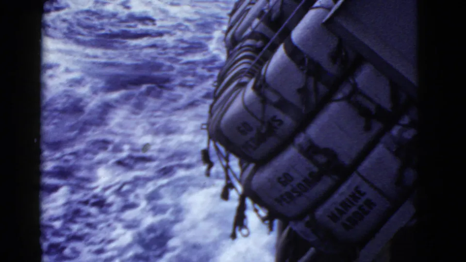 Marked life rafts, including capacity text, sit along the ship edge above a turbulent blue-purple ocean wake.