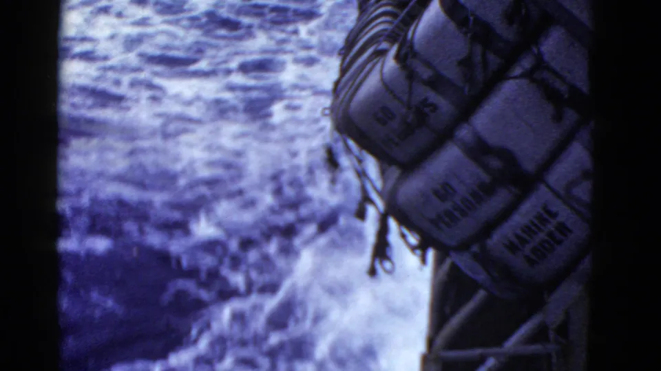 A row of secured ship life rafts, with partial readable markings, hangs above turbulent open water in 8mm footage.