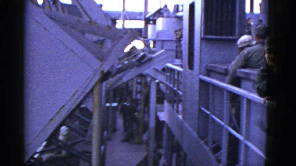 Early-1950s home-movie frame of uniformed men on a vessel deck, with railings, metal structures, and bright sky visible.
