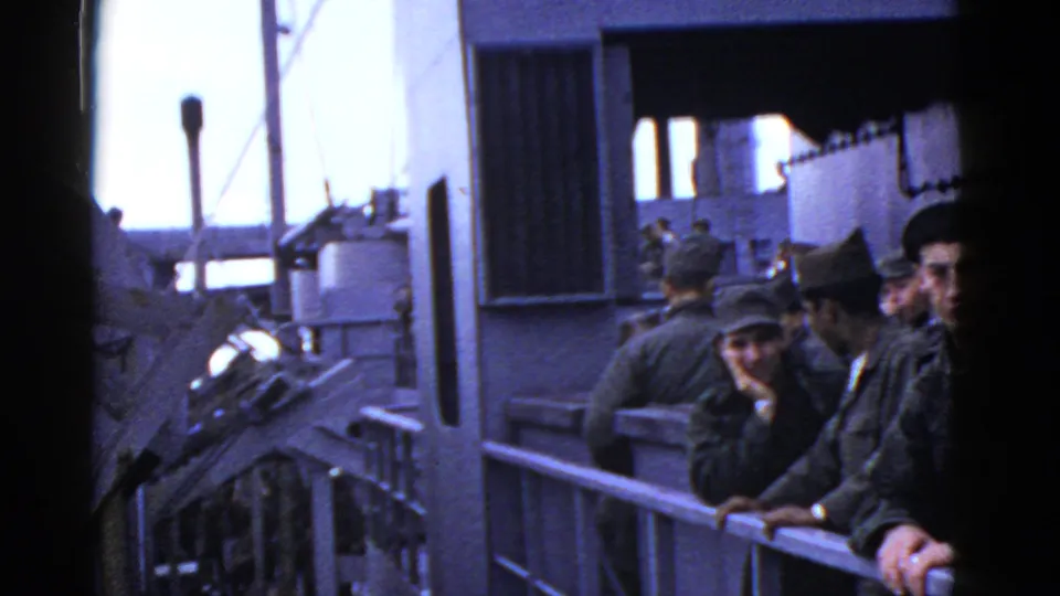 Men in military-style uniforms gather along a vessel railing, framed by dark film edges, metal walls, and bright daylight.