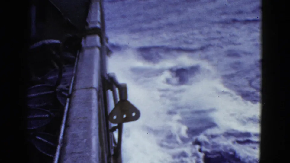 8mm frame from the ship's side, showing railing, dark deck equipment, and white wake moving through open water.