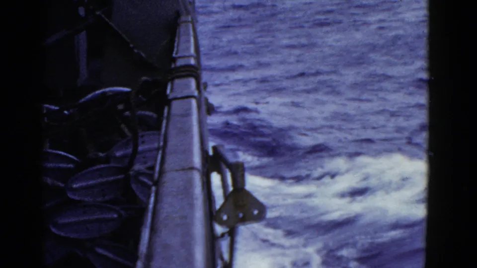 Side-deck view from an 8mm naval home movie, with ship rail, dark equipment, and textured ocean wake filling the frame.