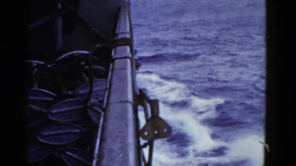 1952 8mm home-movie frame showing foamy water alongside a moving ship, with rail and dark deck hardware at left.