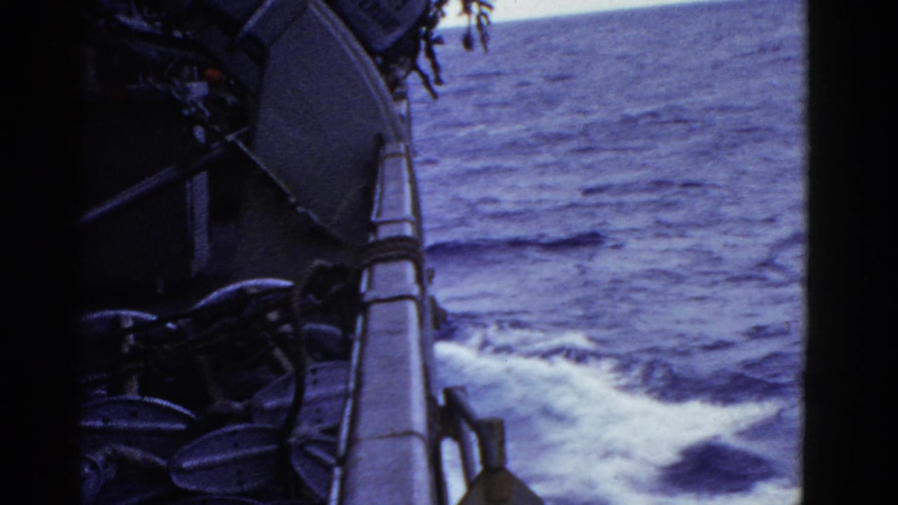 1950s 8mm frame from a ship side deck, with railing and deck equipment overlooking foaming ocean wake.