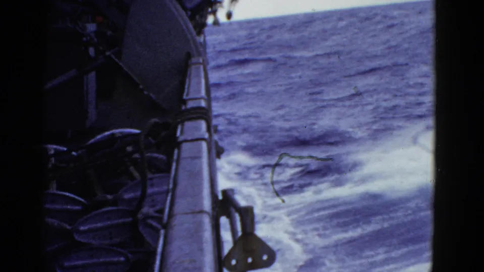 Shipboard 8mm frame with railing, dark deck equipment, and foaming water moving past the vessel.