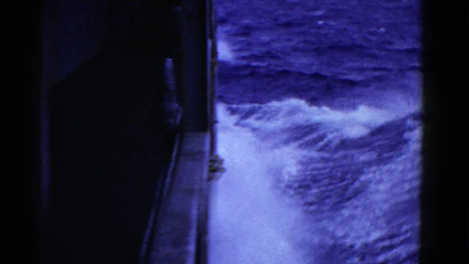 Passenger-view 8mm frame showing the side of a ship, railing, and white wake trailing through blue water.