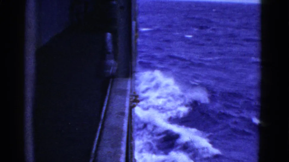1950s home-movie frame looking down along the ship side at churning white wake against deep blue sea.
