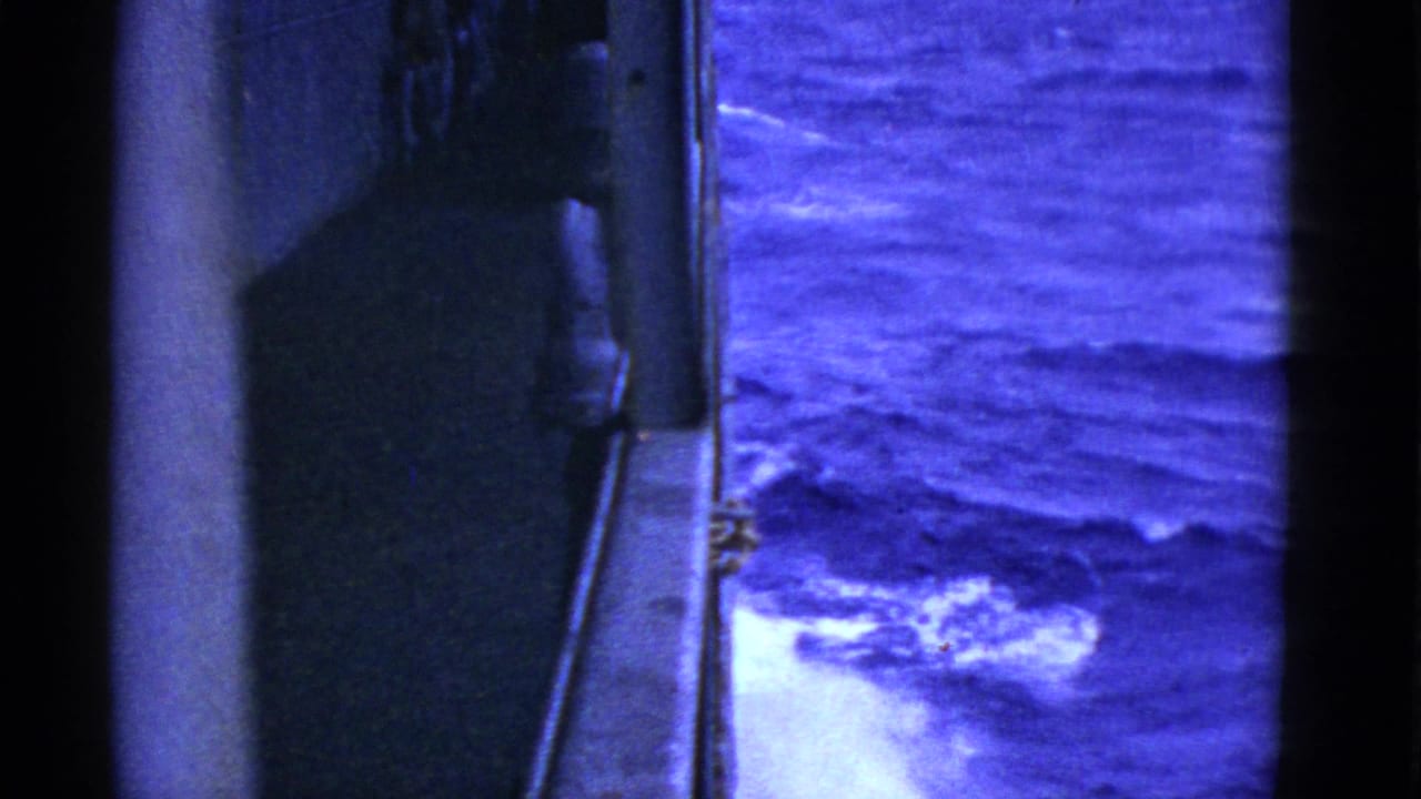 Blue-tinted 8mm shipboard frame showing the vessel edge, railing detail, and foamy rough sea below.