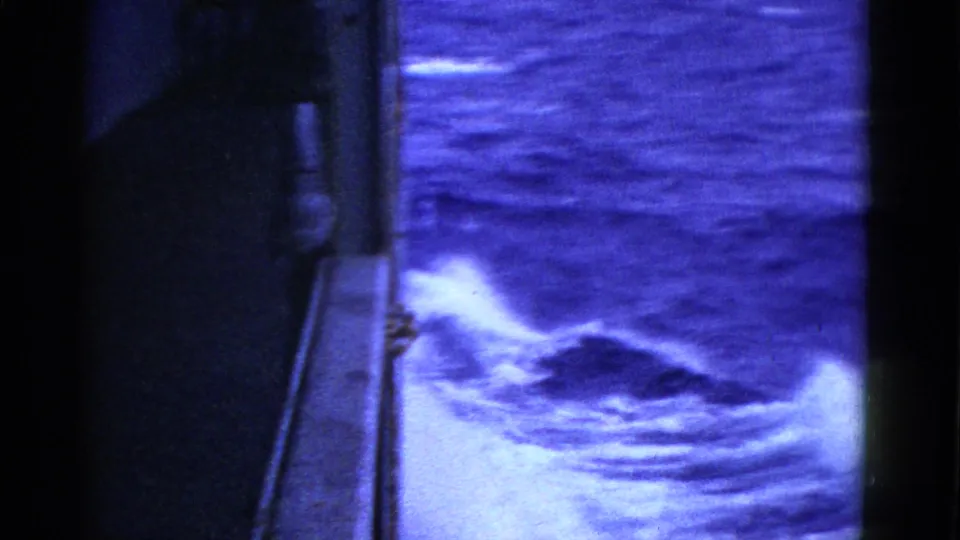 A high shipboard angle shows the dark vessel edge and bright foaming wake in blue-tinted 8mm footage.