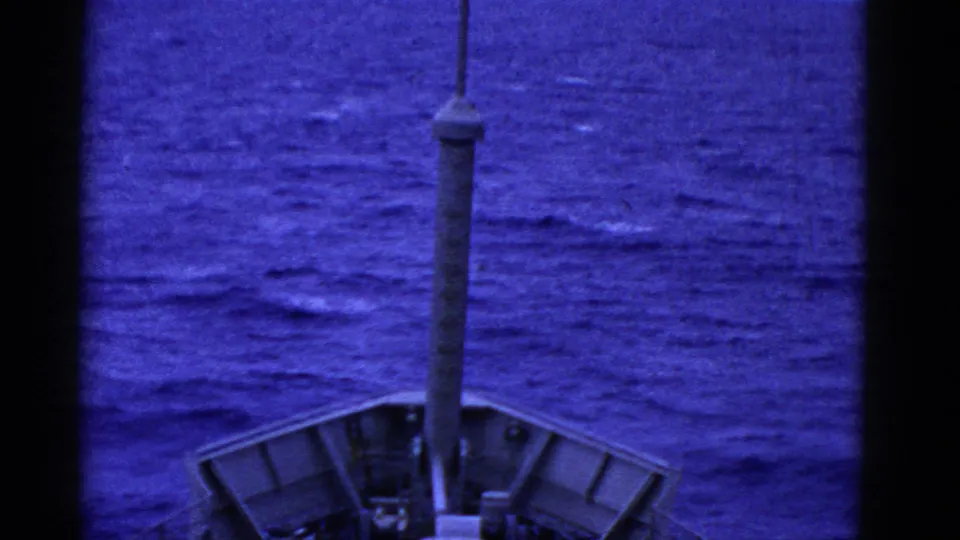 Shipboard home-movie frame showing the bow structure and mast over a blue, grainy ocean surface.