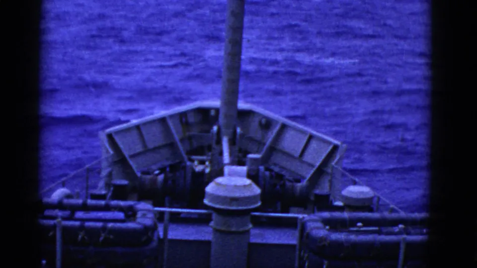 Wider 8mm view from aboard ship, showing bow railings, deck equipment, mast, and blue open water ahead.