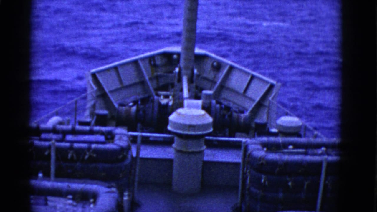 1950s 8mm view of a ship deck at sea, showing stacked life rafts, railings, a central vent, and calm blue water.