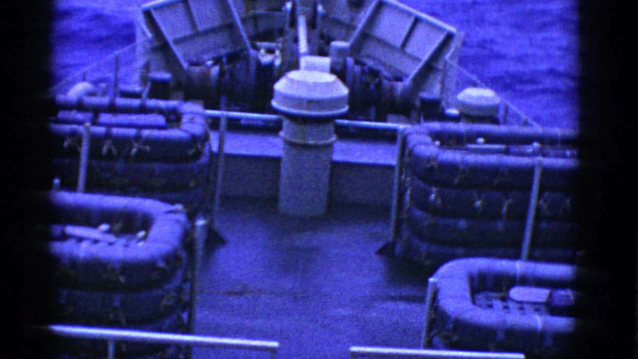 Blue-cast 8mm frame of a ship deck at sea, with life rafts, metal piping, railings, and water visible beyond the bow.