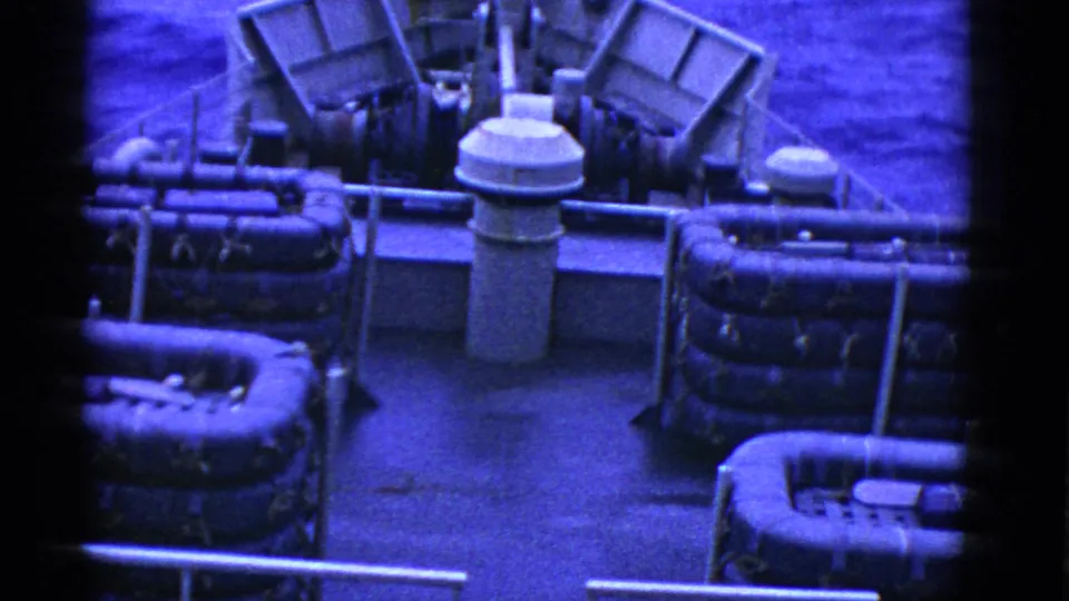Blue-cast 8mm frame of a ship deck at sea, with life rafts, metal piping, railings, and water visible beyond the bow.