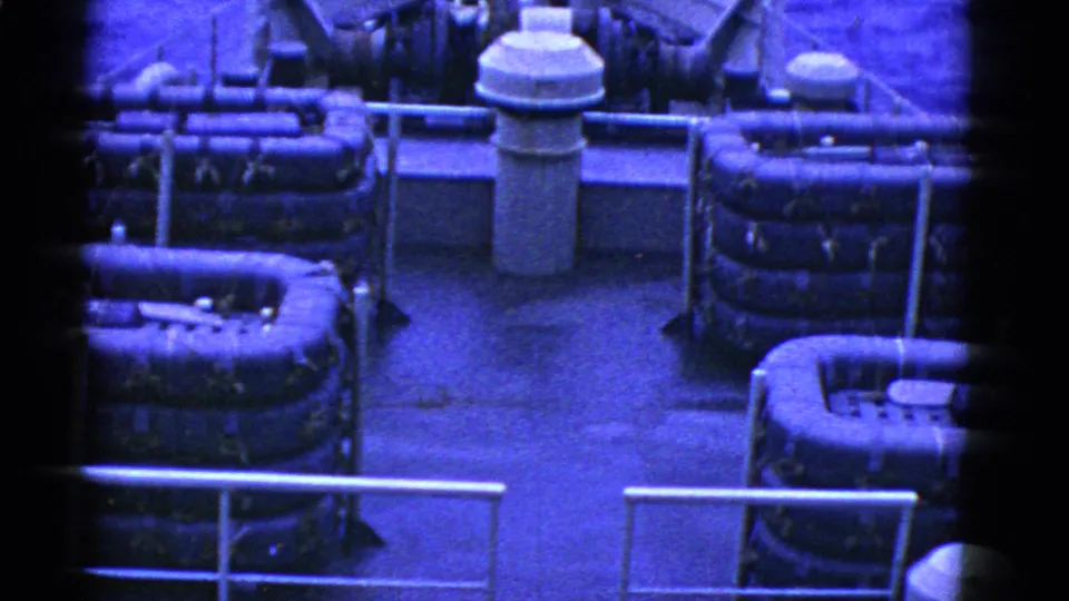 Closer 1950s 8mm view across a ship deck, showing stacked rafts, railings, deck equipment, and strong blue color shift.