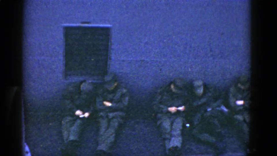 1952 San Francisco 8mm frame of soldiers seated close together against a plain wall, with bags nearby and hands lowered toward personal items.