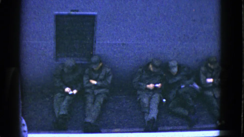 1952 San Francisco home-movie still showing soldiers seated shoulder to shoulder beneath a small dark wall panel, with bags and personal items visible.