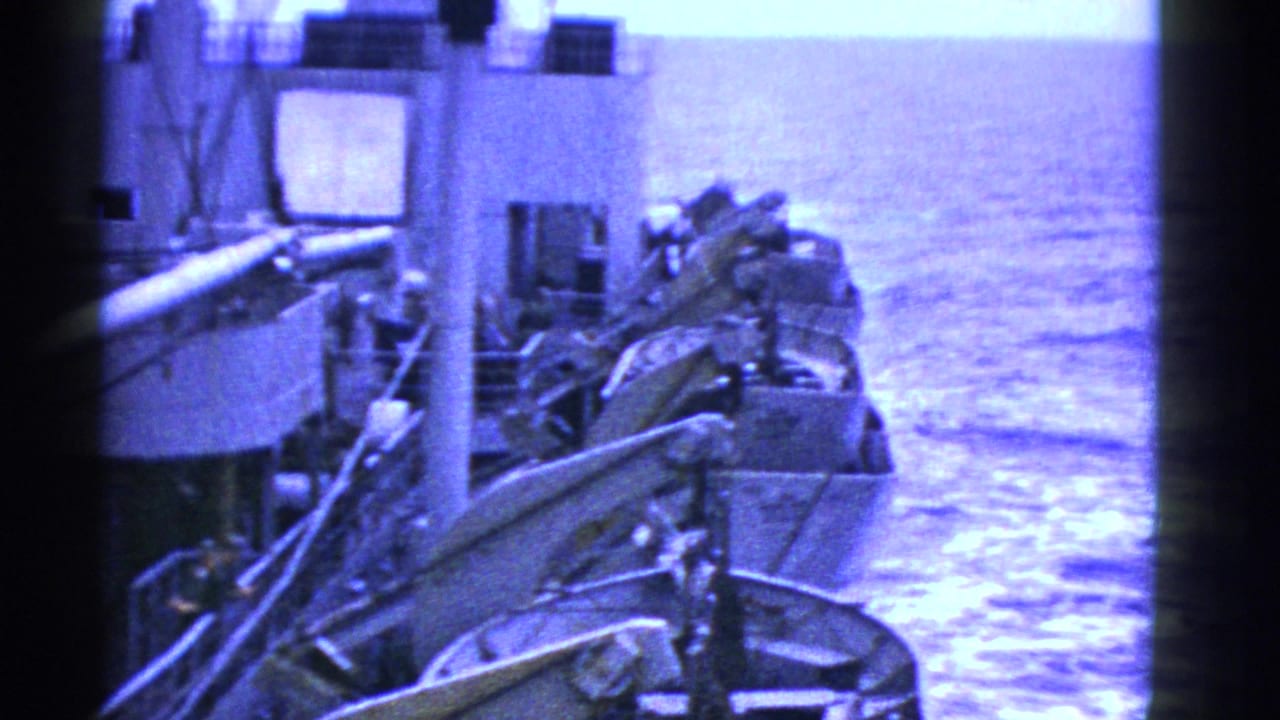 1950s 8mm frame showing lifeboats secured along the side of a ship at sea, with railings and blue water visible.