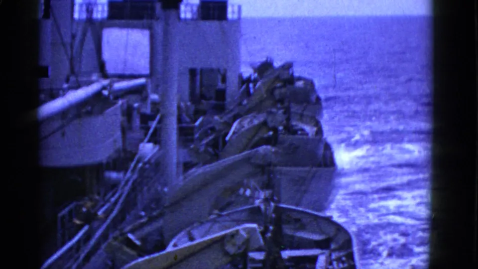 Shipboard view of lifeboats in davits above foaming blue water, with deck structure partly visible.