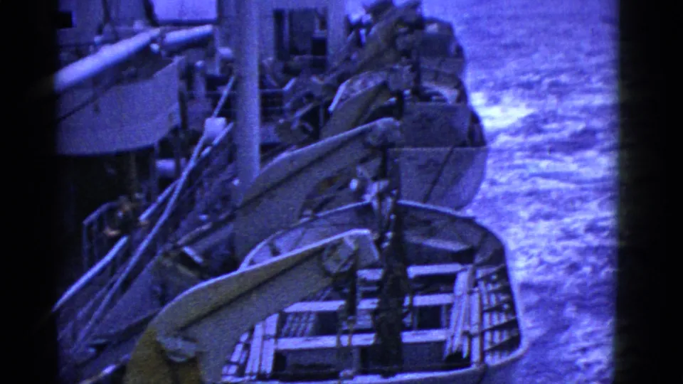 Blue-tinted 8mm view down a ship's side, showing lifeboats, railings, and the sea passing close by.