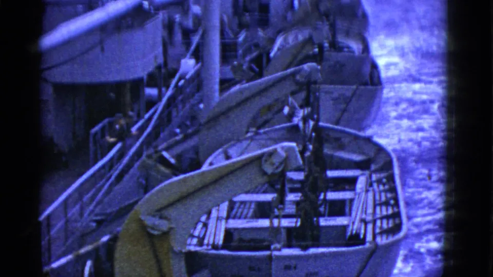 Frame from a 1950s home movie showing multiple lifeboats over the side of a moving ship, with bright wake to the right.