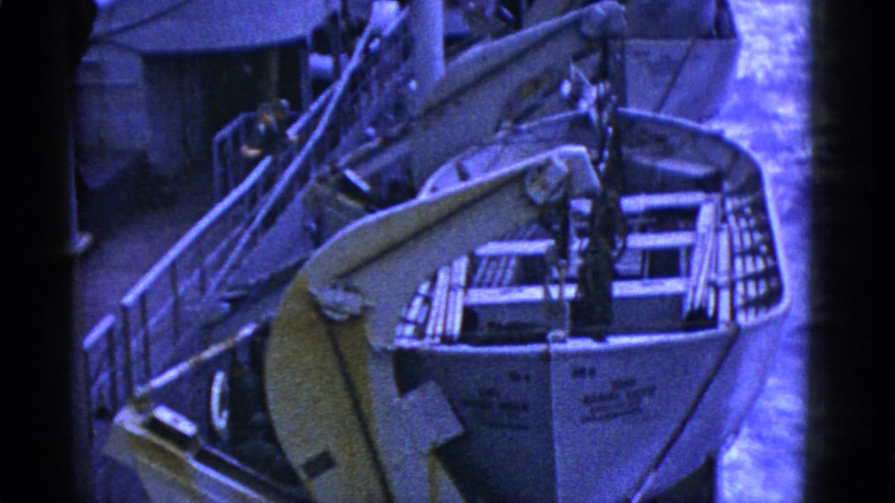 Close view of a rowboat beside a ship with deck gear, railings, rigging, and water beyond.