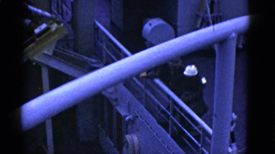 1950s ship deck view with a helmeted worker in dark clothing moving along a metal gangway below railings and pipework.