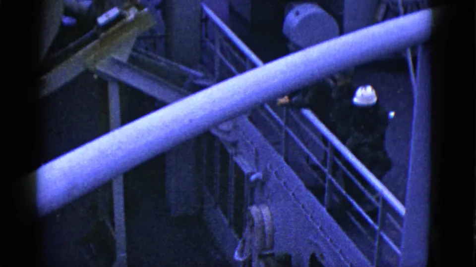 Shipboard 8mm frame showing a worker in a white hard hat beside metal railings, a large pipe, deck fittings, and a narrow gangway.