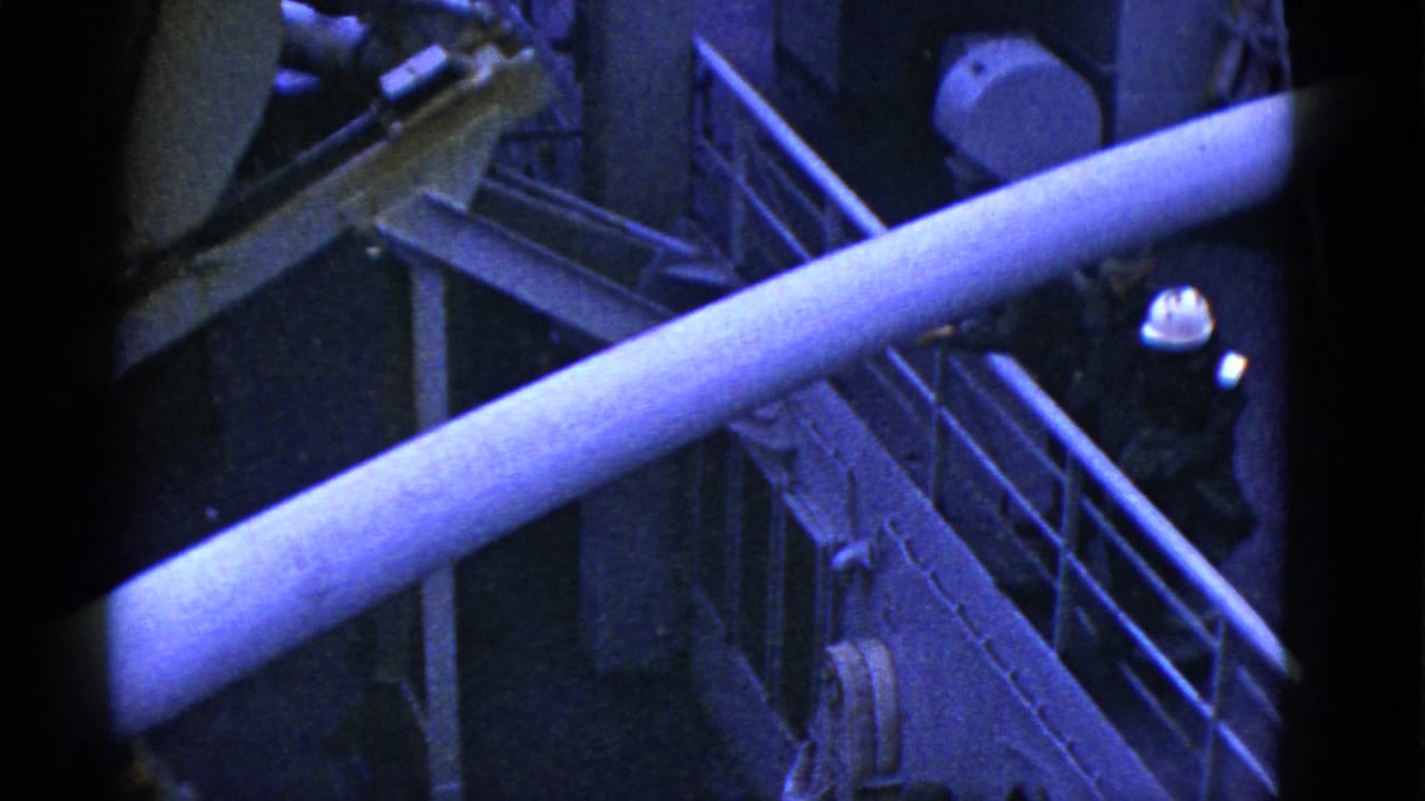 Early 1950s San Francisco 8mm frame looking down on ship deck railings, a crane arm, and a hard-hatted worker near a small boat.