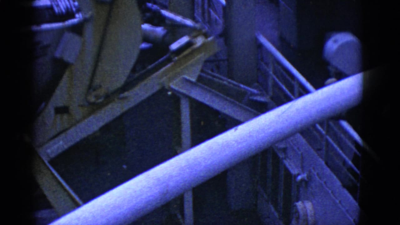 Crane arm and railings cross a dim ship deck with workers partly visible near machinery.